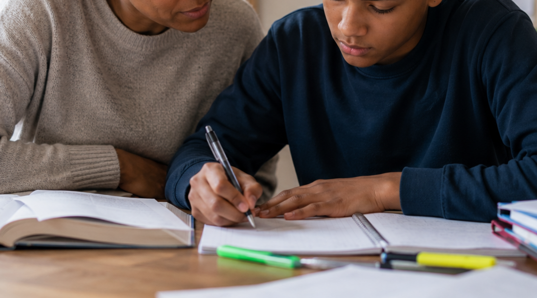 Teen boy studying at a desk beside a supportive parent in a warm, sunlit home environment, surrounded by notebooks and school stationery. Calm, focused study scene showing parent support during schoolwork and learning.