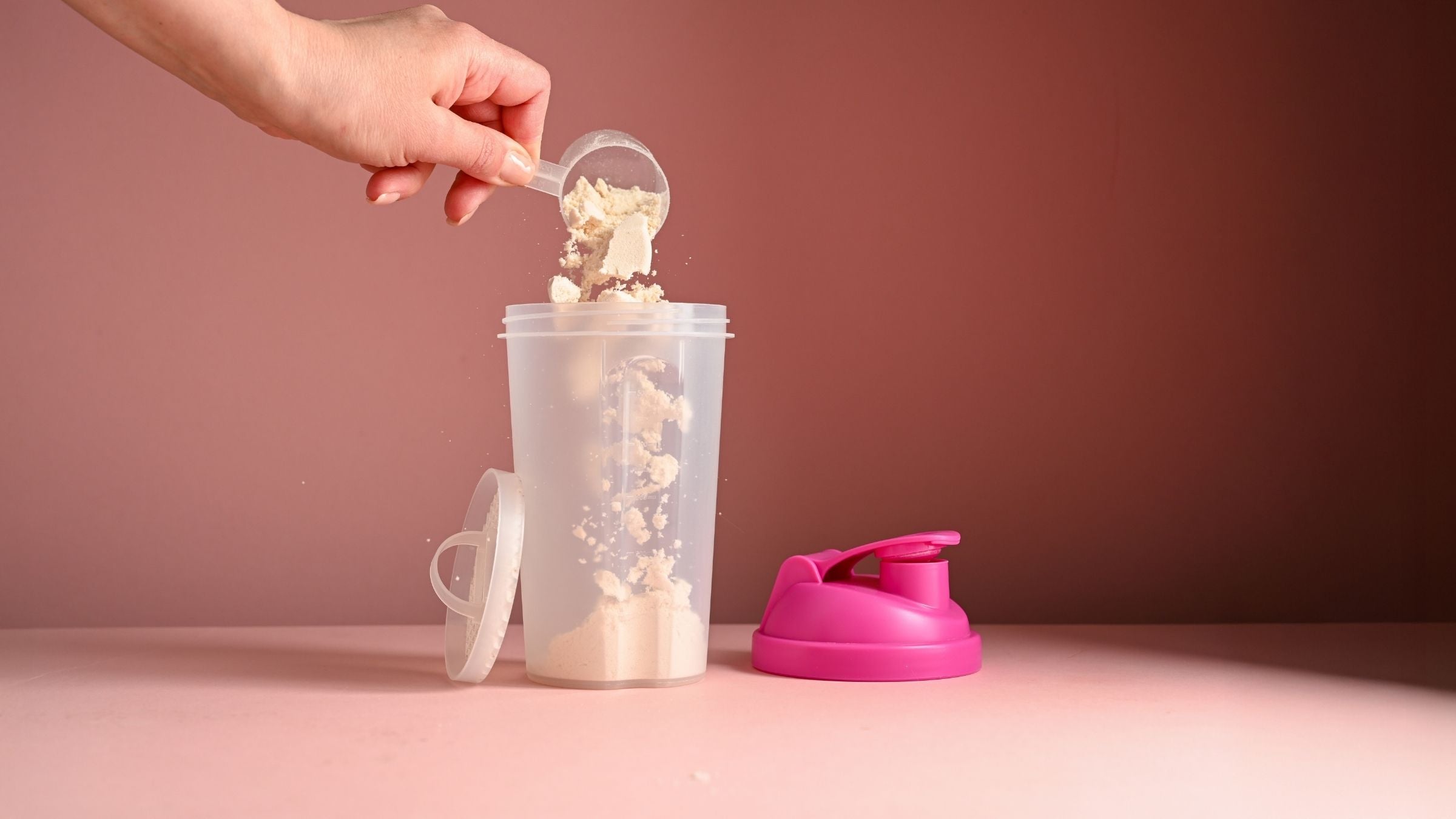 Hand pouring powdered supplement into a transparent shaker bottle with a pink lid against a soft neutral background.