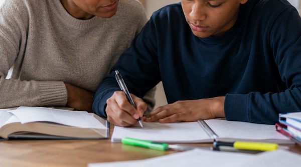 Teen boy studying at a desk beside a supportive parent in a warm, sunlit home environment, surrounded by notebooks and school stationery. Calm, focused study scene showing parent support during schoolwork and learning.